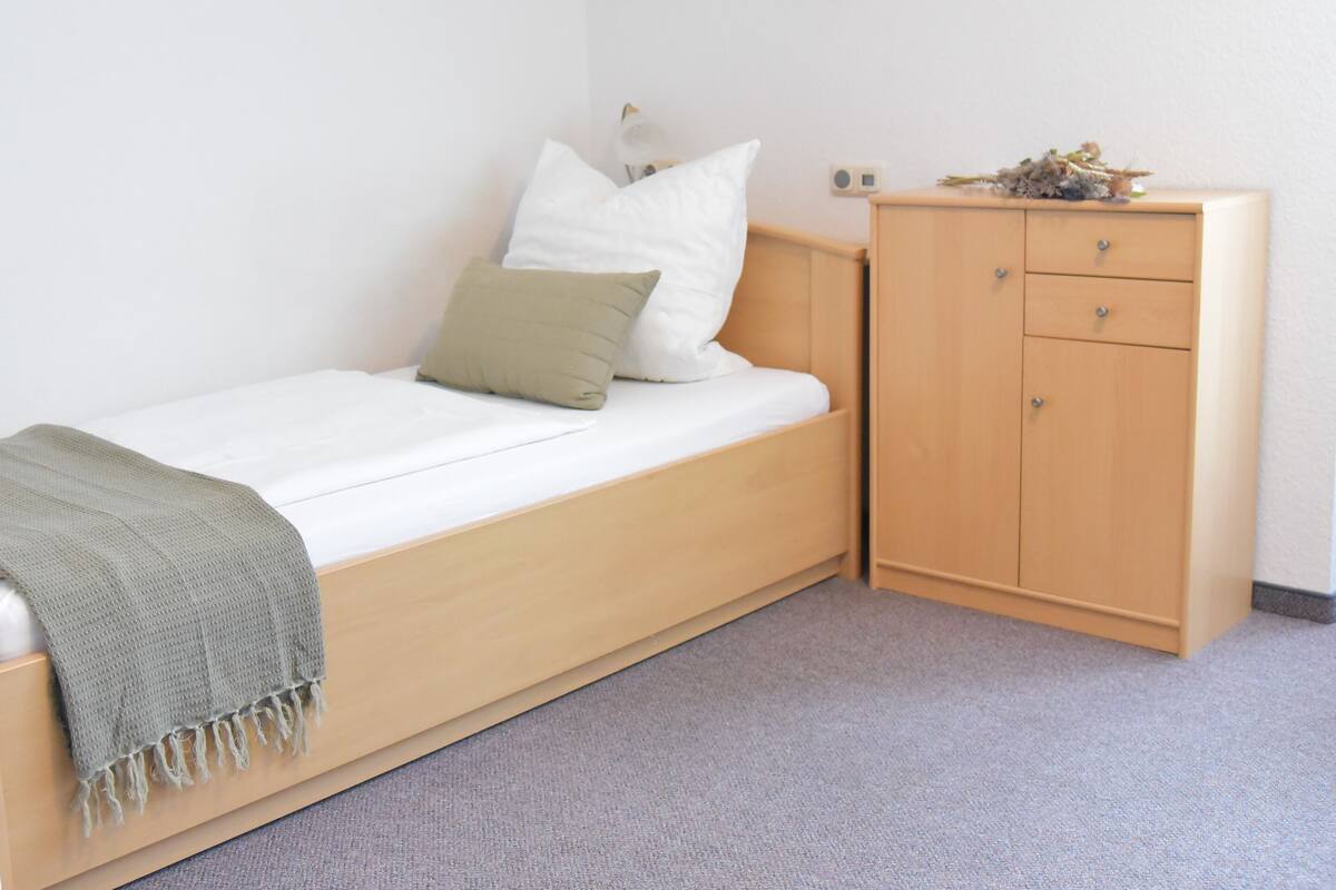 A single-bed room with a wooden bed, a bedside table, a small wardrobe, and a wall-mounted light fixture.