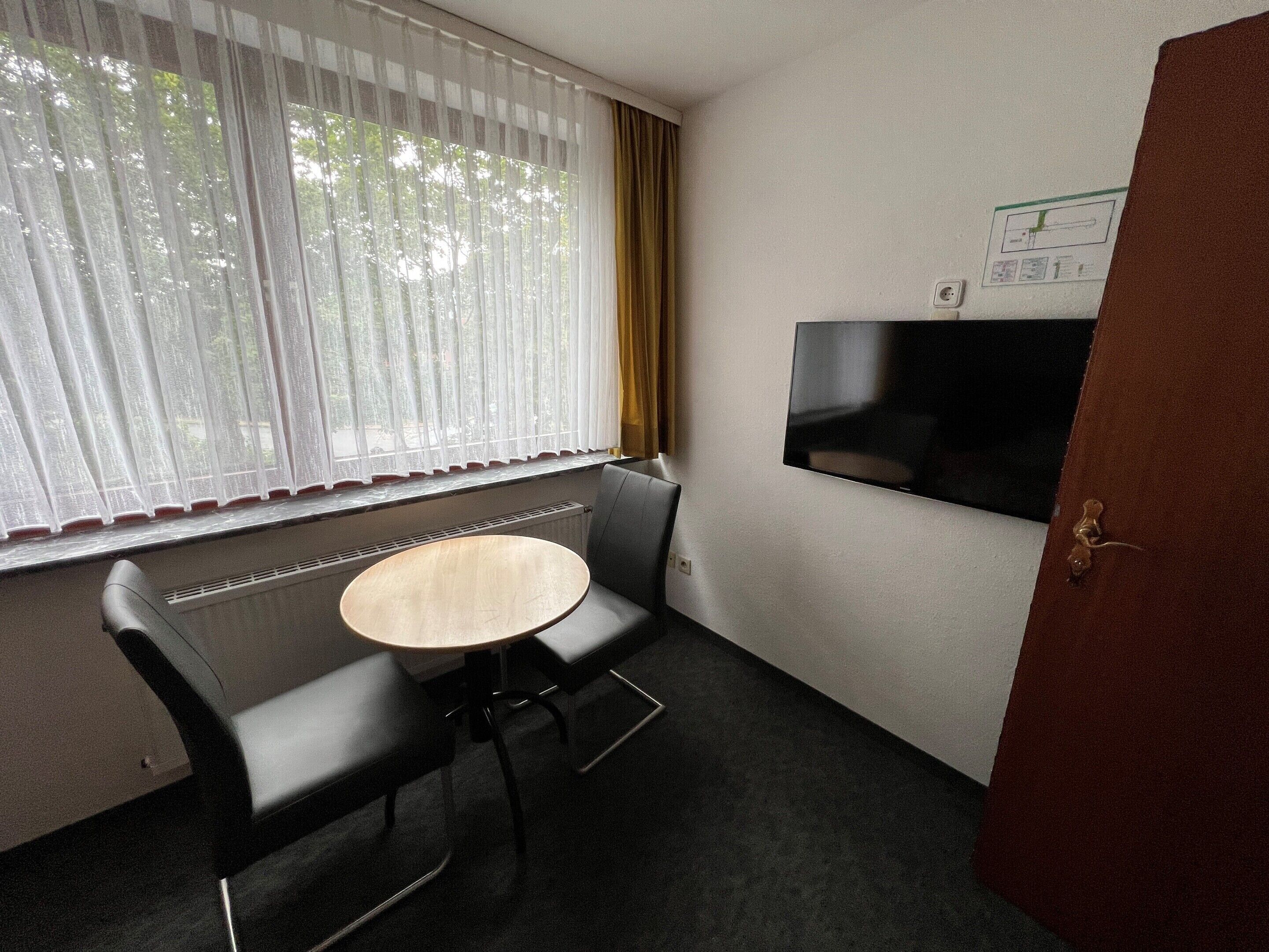 A small room with a single sofa, a round table, a TV mounted on the wall, and a window with sheer curtains.