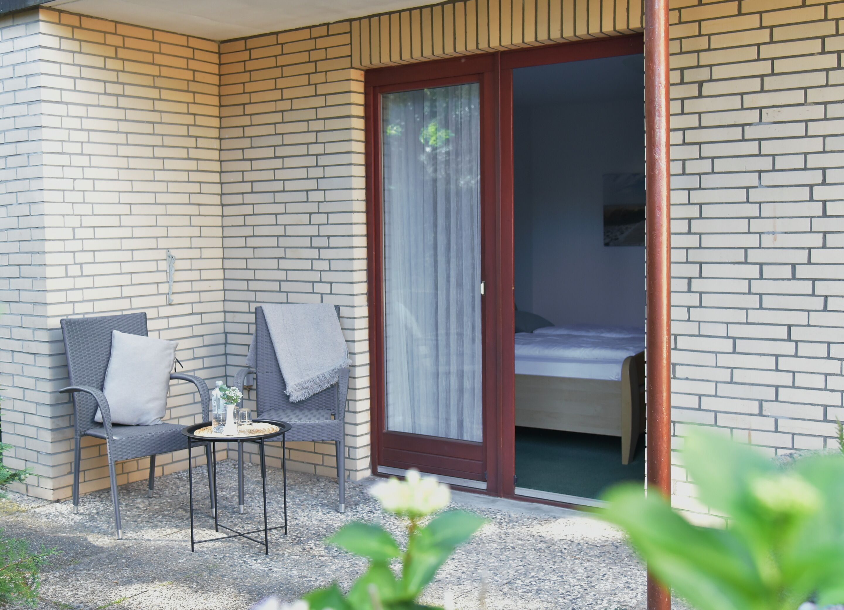 Een klein terras met twee stoelen en een tafel, een schuifdeur naar een slaapkamer, en een bakstenen muur.
