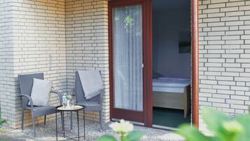 Een klein terras met twee stoelen en een tafel, een schuifdeur naar een slaapkamer, en een bakstenen muur.