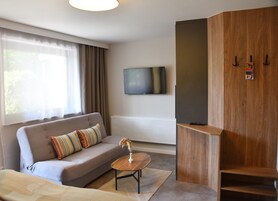 A modern hotel room with a grey sofa, a round wooden coffee table, a TV mounted on the wall, and a wooden wardrobe with hooks.