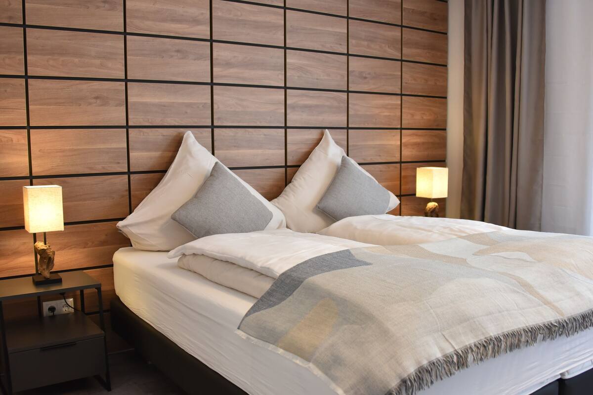 A neatly made bed with white linens and grey pillows, a bedside table with a lamp, and a wooden panel wall.