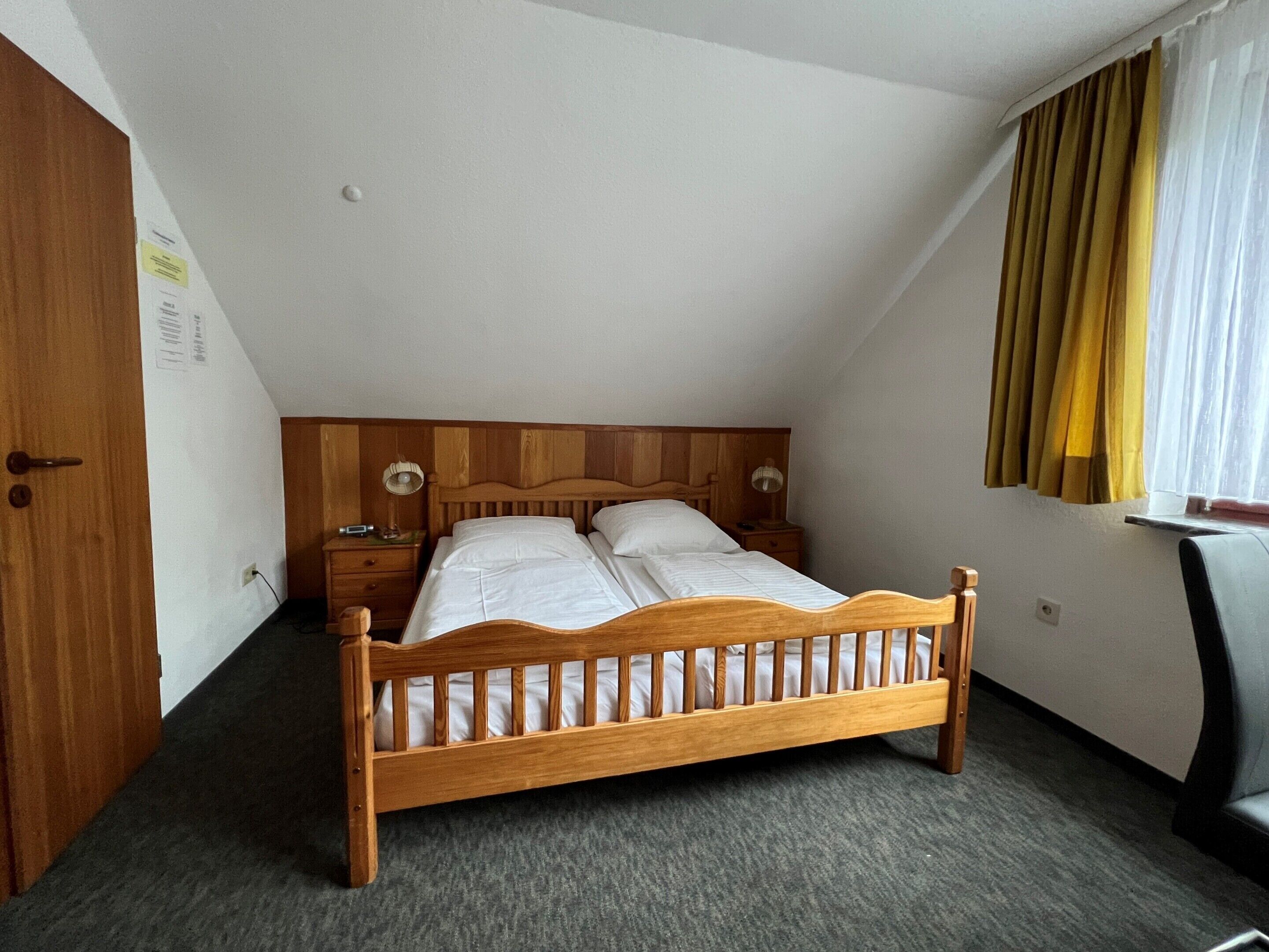 A single bed in a room with a sloped ceiling, a wooden headboard, a bedside table with a lamp, and a window with curtains.