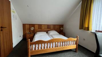 Una cama individual en habitación con techo inclinado, cabecera de madera, mesita de noche con lámpara y ventana con cortinas.