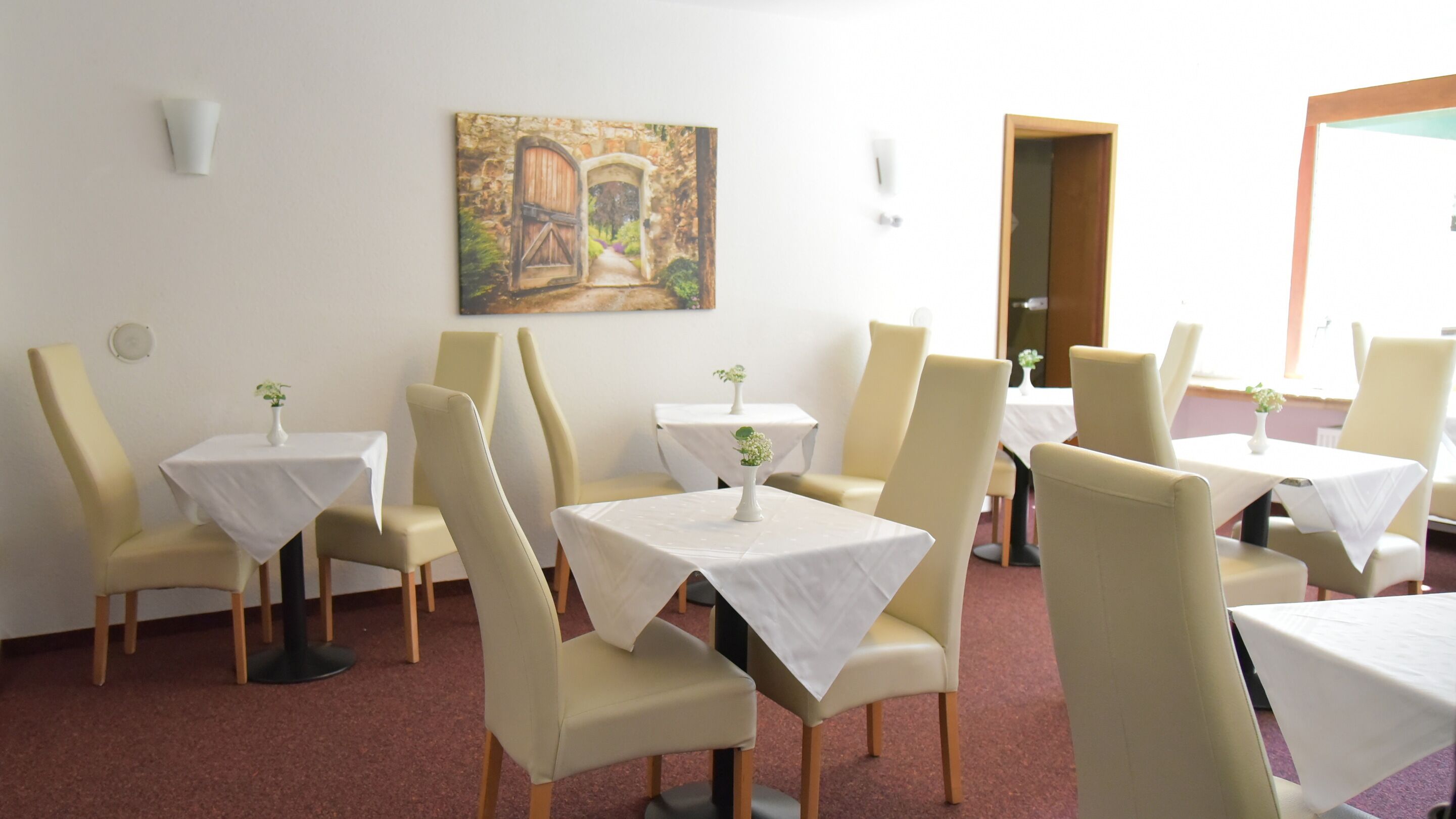 Een eetruimte met tafels en stoelen, een schilderij aan de muur en een deur die naar buiten leidt.
