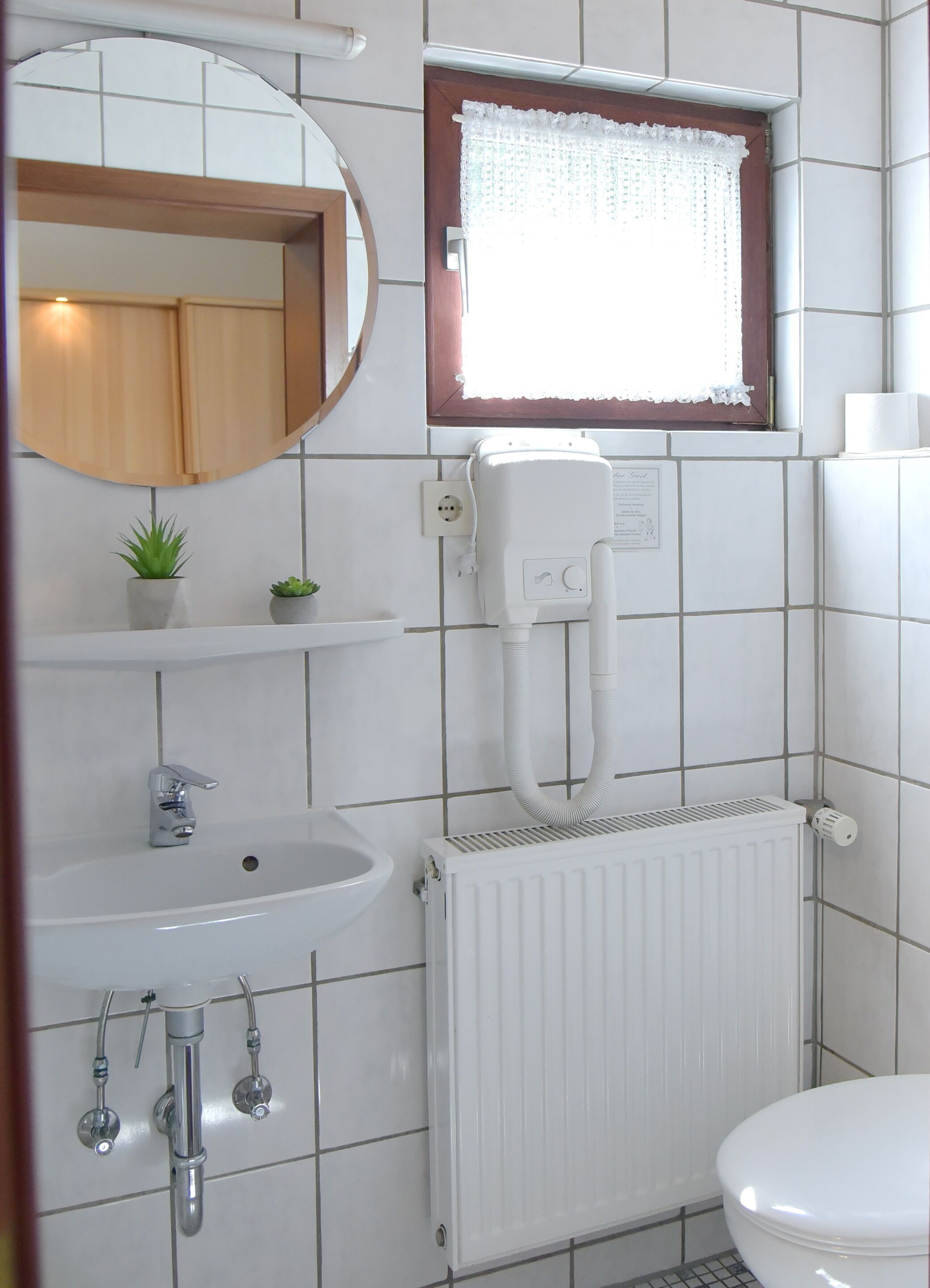 Une salle de bain avec un lavabo blanc, un miroir, une fenêtre ornée de rideaux en dentelle, un radiateur et des toilettes.