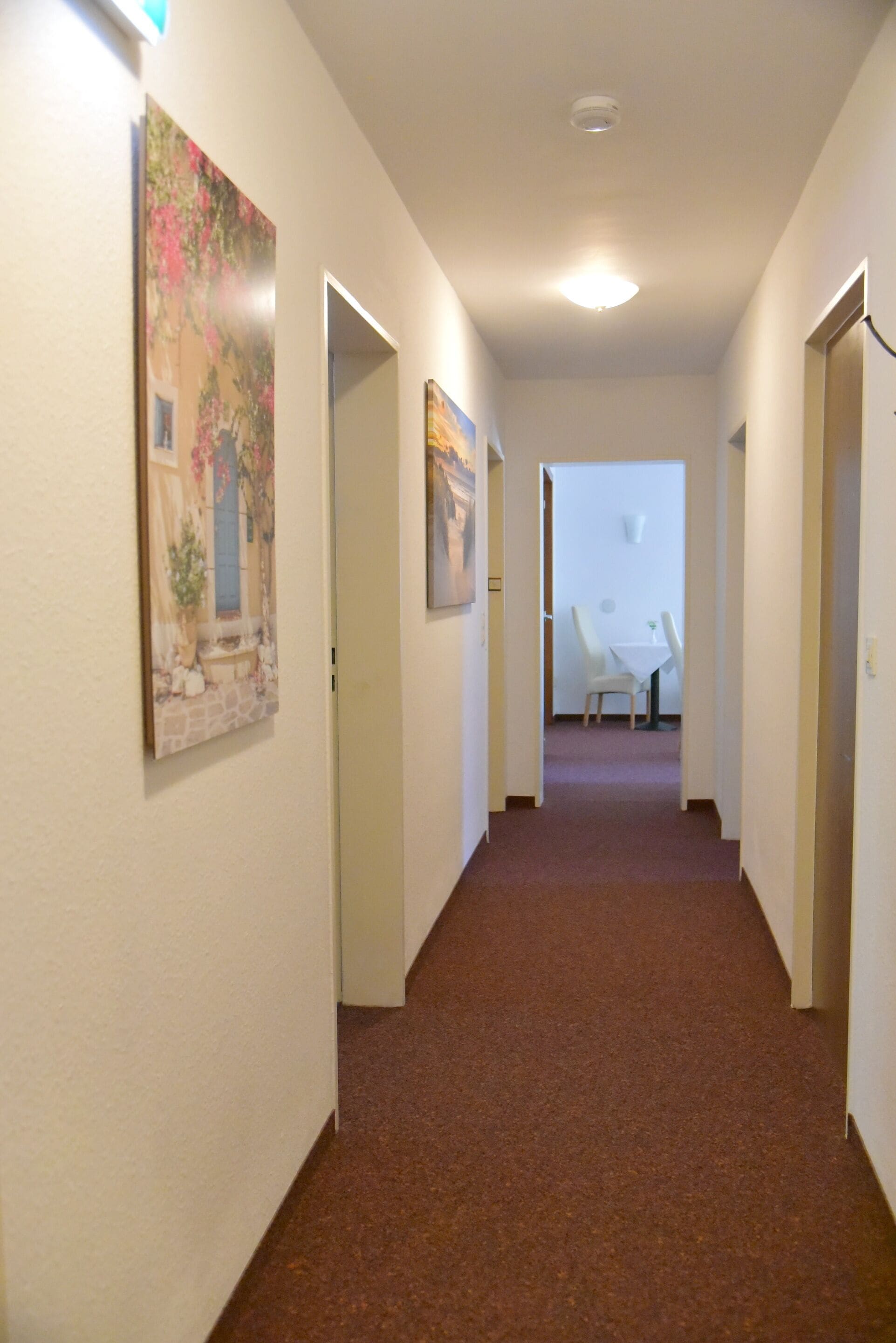 Ein Flur mit Teppichboden, weißen Wänden und gerahmten Kunstwerken. Rechts befinden sich Türen und in der Ferne ein Tisch mit Stühlen.