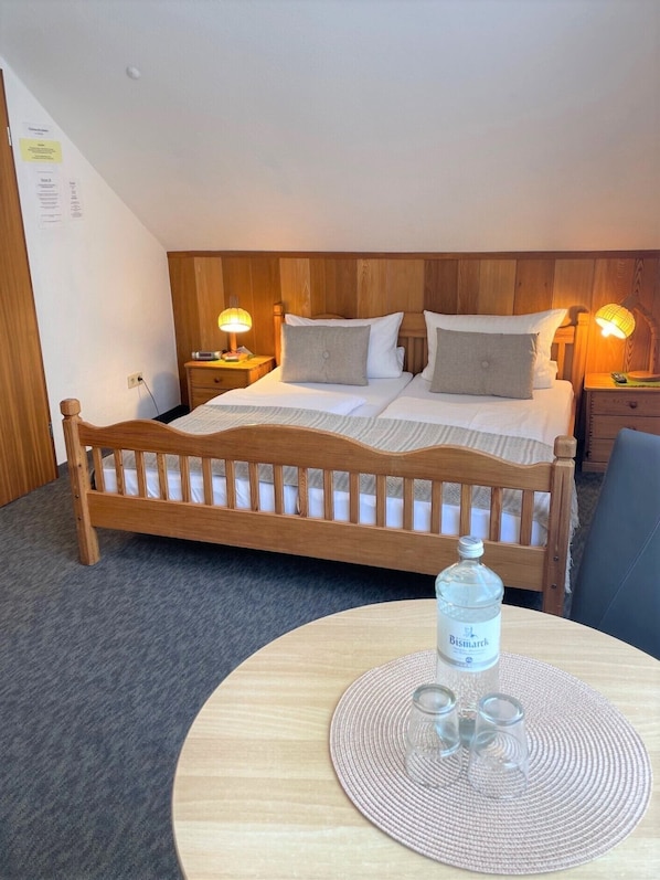A hotel room with a wooden bed, two bedside tables with lamps, a round table with a bottle of water and glasses, and a grey carpet. - Pension Haus Bambi (Moelln)