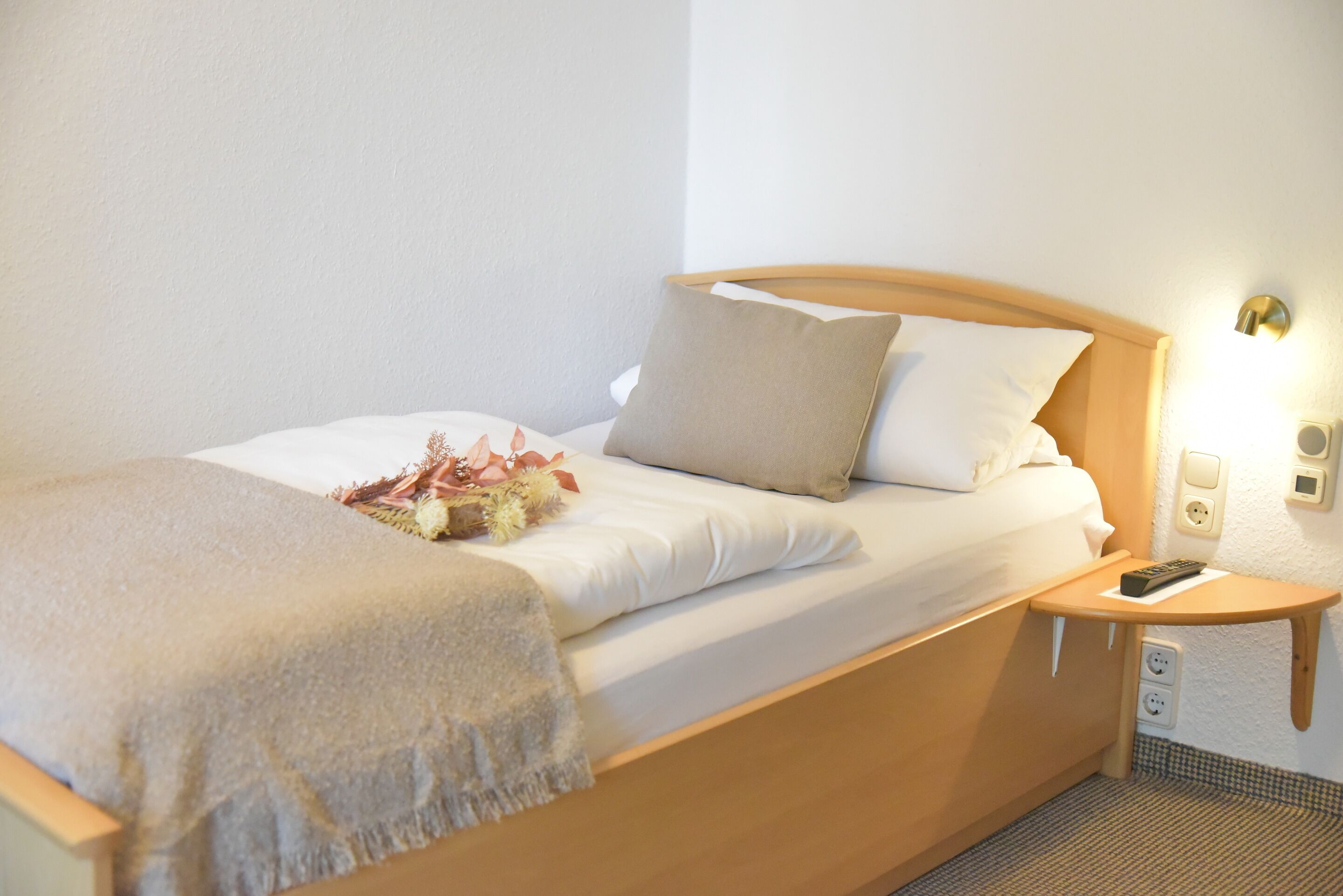 A neatly made bed with a wooden headboard, a bedside table with a remote control, and a wall socket.