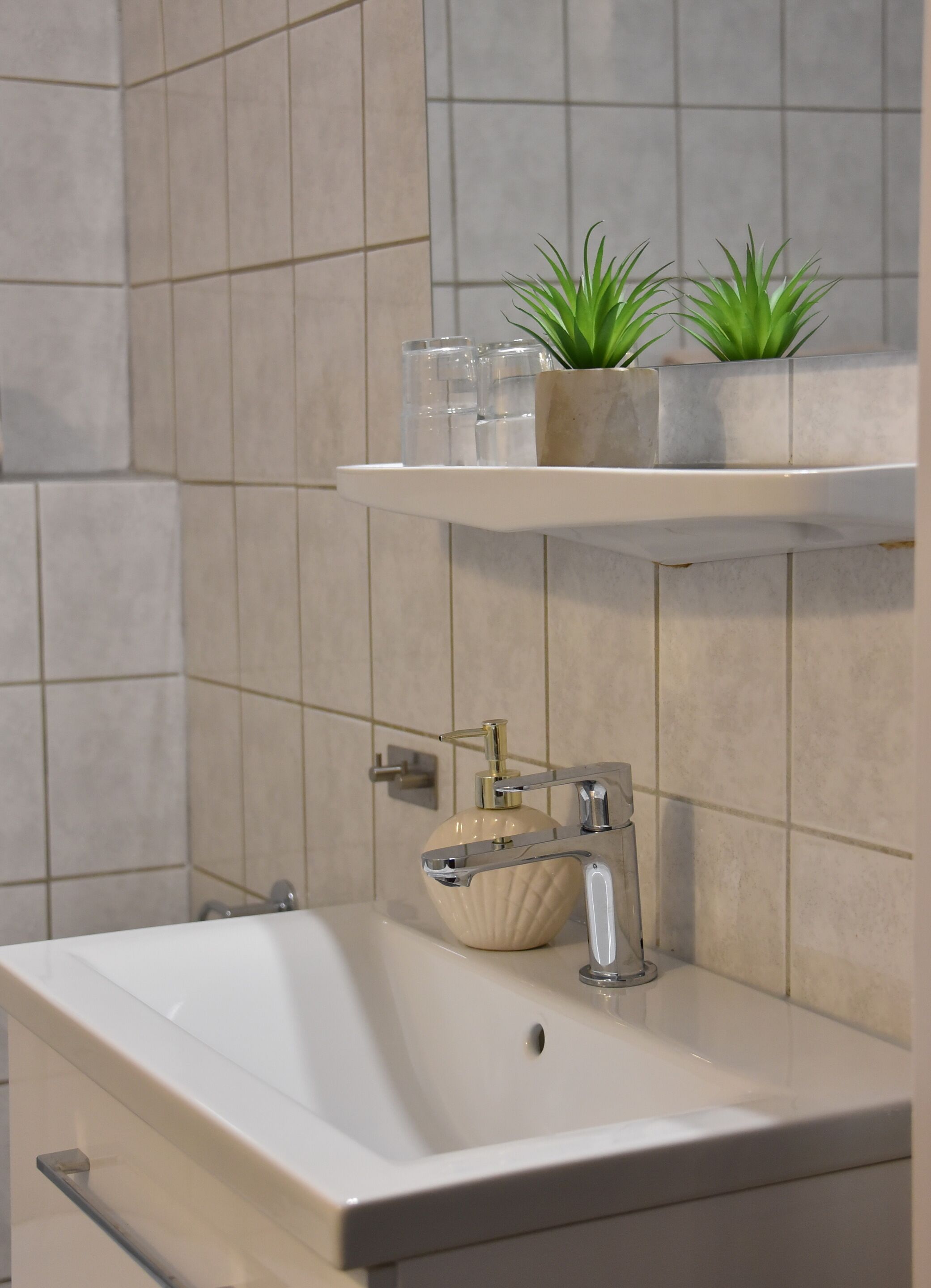 Un baño con un lavamanos blanco, un dispensador de jabón color beige y dos plantas en macetas sobre una repisa.