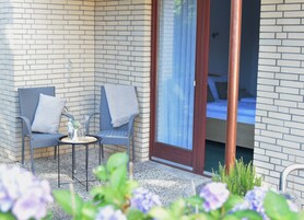 A small outdoor seating area with two chairs and a table, a sliding glass door leading to a bedroom, and a garden with flowers.