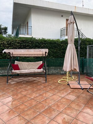 Une terrasse avec un fauteuil suspendu, une table couverte et un porte-parapluie.