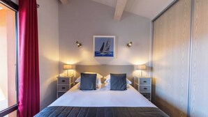 A hotel room with a bed, two bedside tables with lamps, a painting of a sailboat on the wall, and a window with red curtains. - Appartements Hôtel & Spa Le Madeloc Collioure Centre (Collioure)