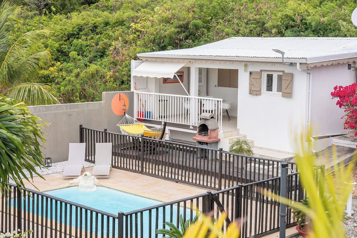 A white house with a balcony, a swimming pool, and lush greenery in the background.