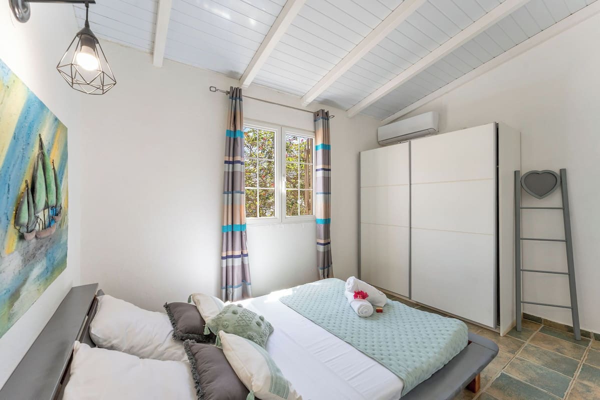 A bedroom with a bed, pillows, a painting, a window with curtains, an air conditioning unit, and a ladder.