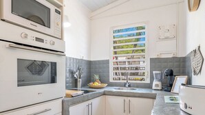 A compact kitchen with white cabinets, a built-in oven, a window with a view, and a countertop with a blender and a bowl of fruit. - CASA LYDIE - ÉVASION CRÉOLE AVEC PISCINE (Saint-François)