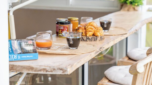 A breakfast table with croissants, juice, and coffee, set on a wooden deck with chairs and a view of palm trees. - CASA LYDIE - SUITE PISCINE PRIVÉE (Saint-François)