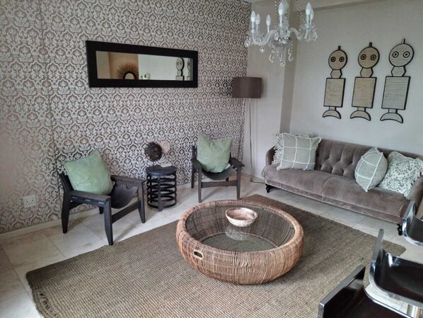 Ein Wohnzimmer mit gemustertem Wandpapier, einem runden Korbstuhl-Couchtisch, einer Couch, zwei Sesseln, einer Stehlampe und Wandbildern.