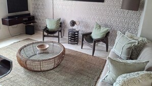 Ein Wohnzimmer mit einem runden Korbtisch, zwei Sesseln, einem Sofa, einem Fernseher, einem Spiegel und ornamental gestalteter Tapete.