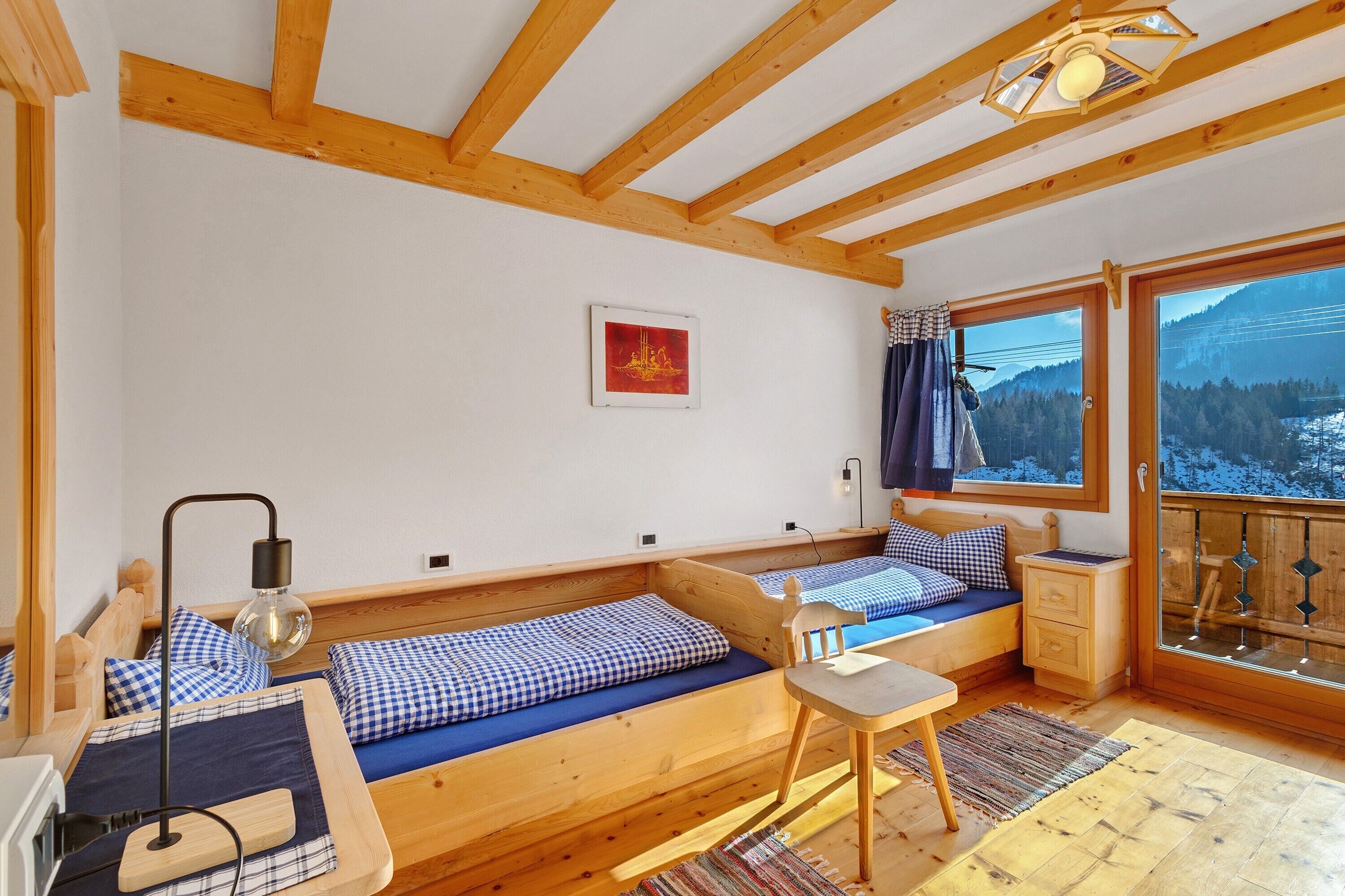 Une cabane avec des poutres en bois, un lit avec une couette à carreaux, un bureau et une fenêtre donnant sur la montagne.