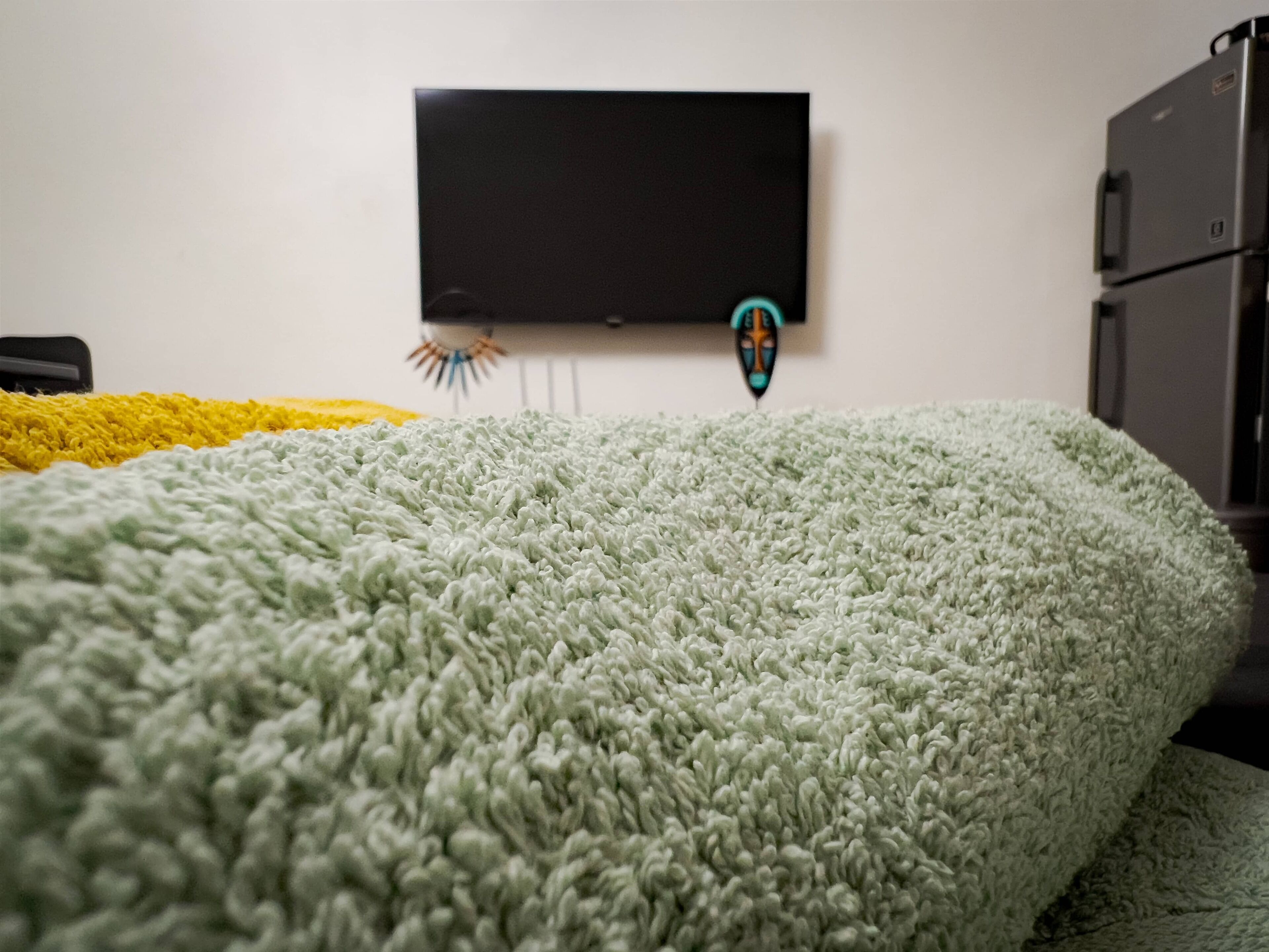 A room with a bed covered in a green blanket, a flat-screen TV mounted on the wall, and a small decorative item hanging below it.