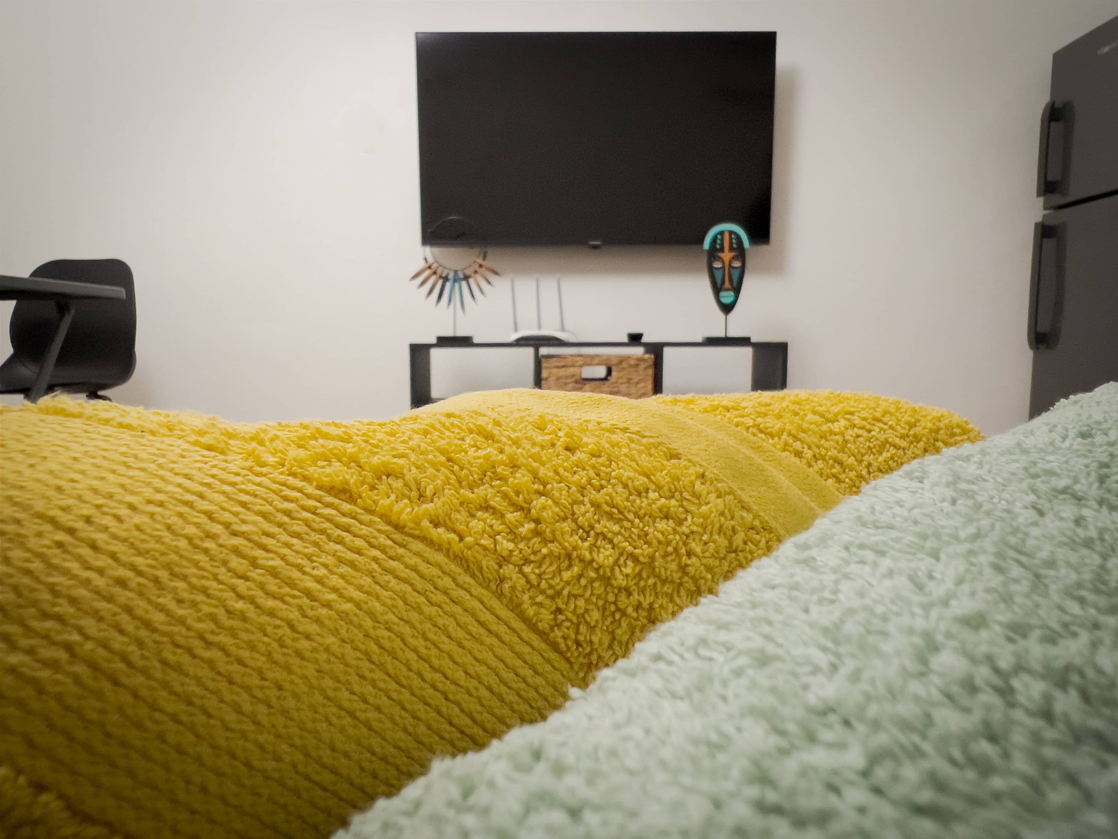 A bed with a yellow and white blanket, a flat-screen TV mounted on the wall, and a small wooden cabinet below the TV.