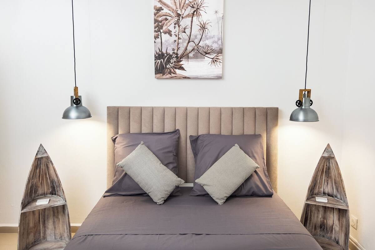 Un dormitorio con una cama, dos almohadas, un cuadro en la pared y dos lámparas colgantes.