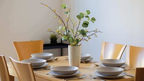 Un tavolo da pranzo apparecchiato con piatti, ciotole e posate bianche, decorato al centro con un vaso di verde fogliame e fiori gialli.