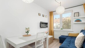 A modern living room with a white dining table, two white chairs, a blue sofa, and a TV mounted on the wall. - Comfortable Apartment | Close to Old Town and Place to Work (Gdańsk)