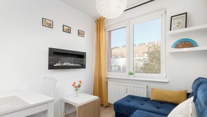 A modern living room with a sofa, a small table, a white chair, a window with yellow curtains, and wall-mounted shelves with decorative items. - Comfortable Apartment | Close to Old Town and Place to Work (Gdańsk)