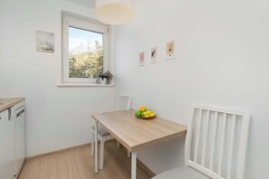 Et lille, rent værelse med et træbord, hvide stole, et vindue med udsigt til træer og en radiator.