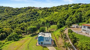 A house surrounded by lush greenery, with a clear sky and a hilly landscape in the background. - Oceanview Luxury Villa Near El Yunque w/Hot Tub (Las Coles)