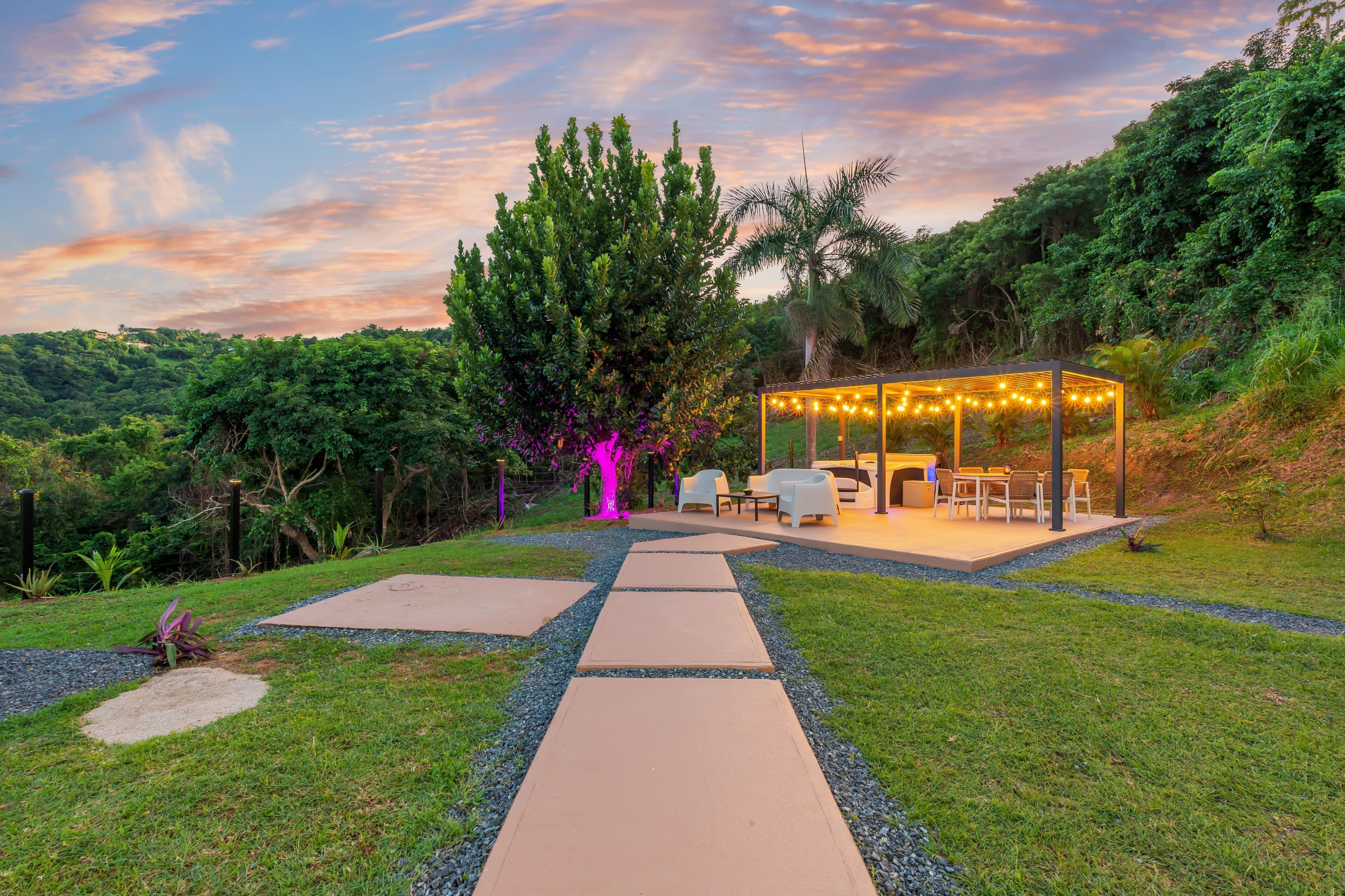 A covered outdoor seating area with a table and chairs, surrounded by a garden with a pathway leading to it.