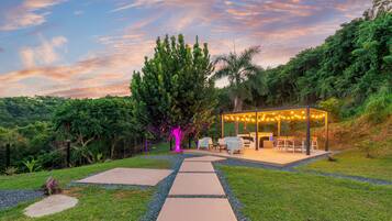 A covered outdoor seating area with a table and chairs, surrounded by a garden with a pathway leading to it.