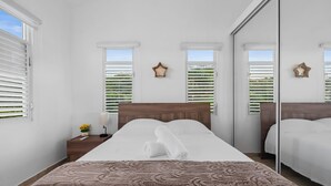 A neatly arranged bedroom with a large bed, white walls, and a wooden headboard. There are two windows with white blinds, and a small nightstand with a lamp and a vase of flowers. - Oceanview Luxury Villa Near El Yunque w/Hot Tub (Las Coles)