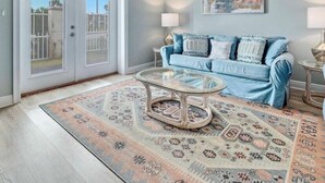 Ein Wohnzimmer mit Deckenventilator, einem blauen Sofa, einem Glastisch, gemusterten Teppichen und einer zur Balkontür führenden Tür.