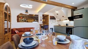 Un coin repas avec une table en bois dressée pour le repas, une cuisine en arrière-plan et un espace de vie chaleureux avec un canapé et une cheminée.