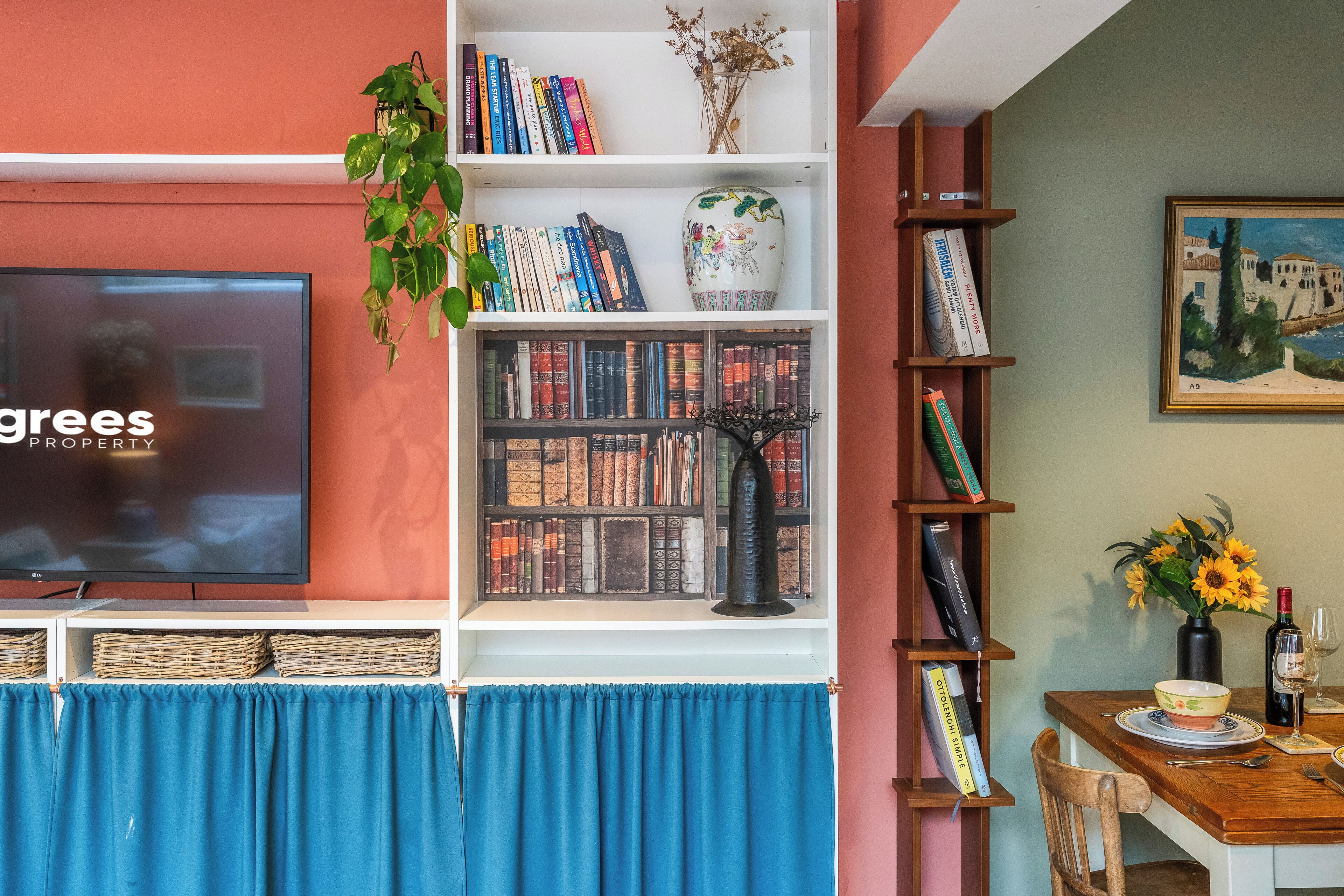 A living room with a television, bookshelves, a dining table with a vase of sunflowers, and a painting on the wall.