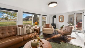 A modern living room with a leather sofa, a beige armchair, and a wooden coffee table. There is a fireplace, a large window, and a view of the outdoors. - Sunset Haven Dog-friendly Home Near Trails/lake (Fort Collins)