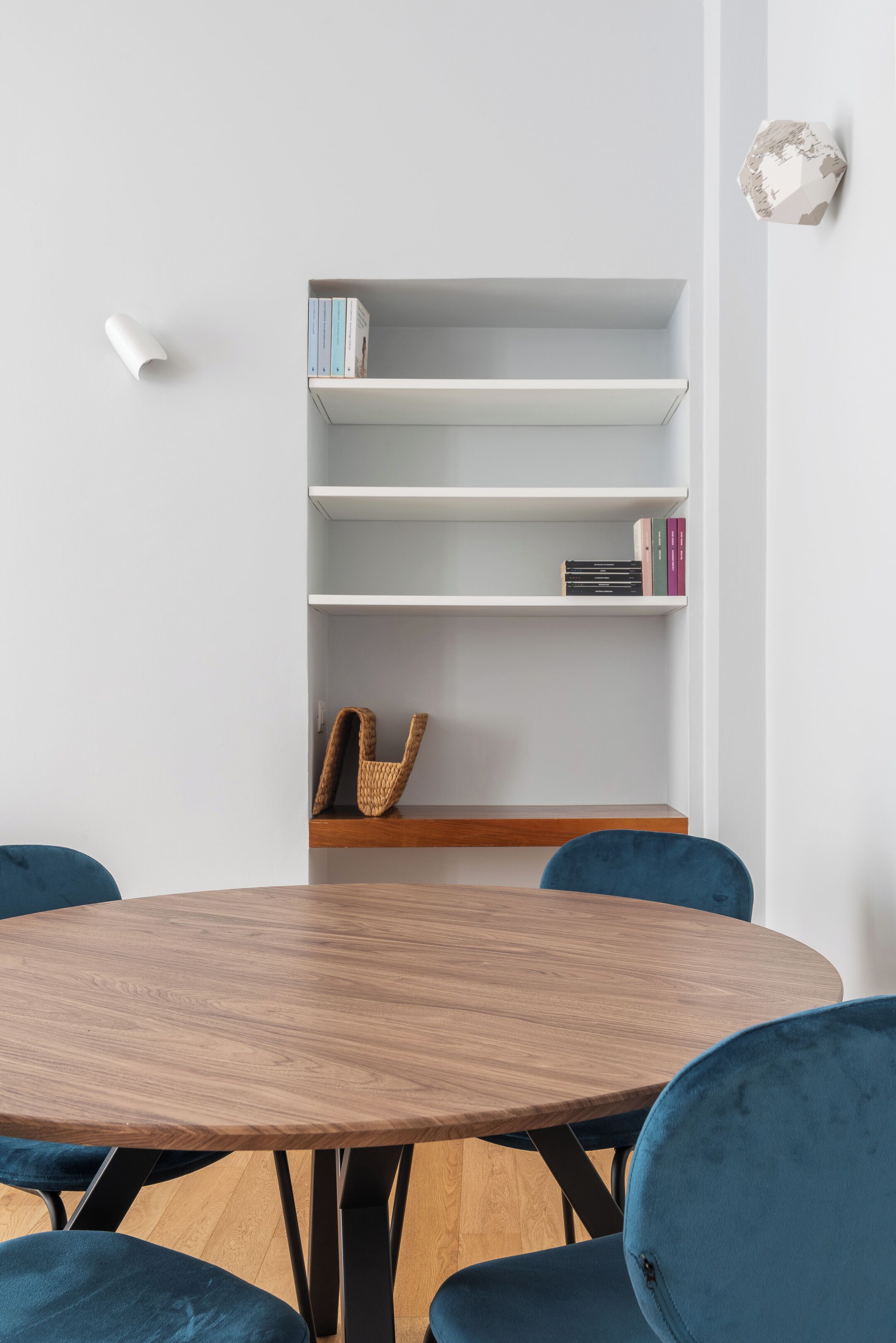 A round wooden table with four blue chairs, a white bookshelf with books and a woven basket, and a decorative wall piece.