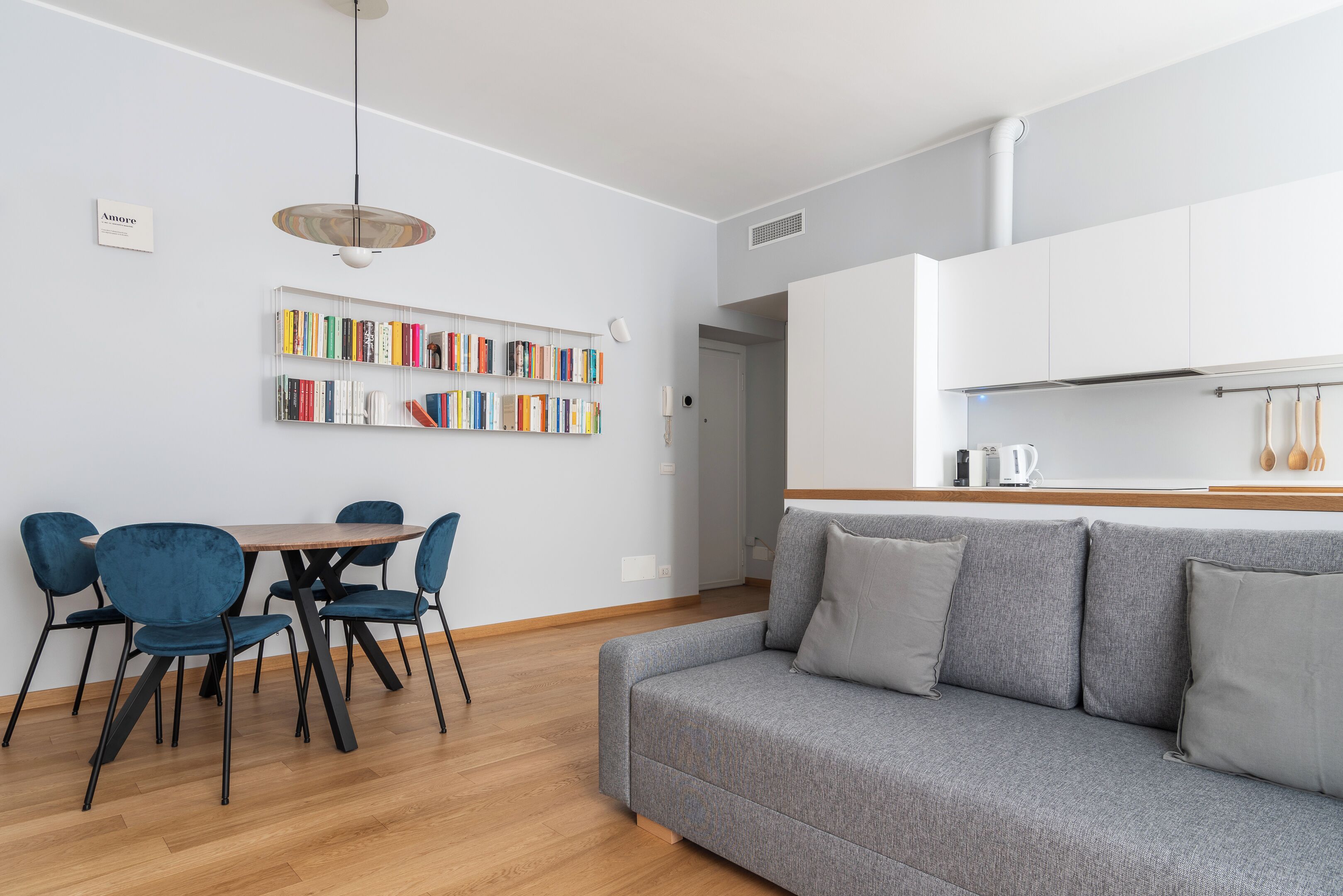 A modern living room with a grey sofa, blue chairs, a wooden dining table, and a bookshelf with colorful books.