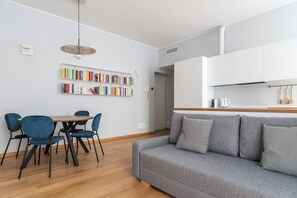 Ein modernes Wohnzimmer mit einem grauen Sofa, blauen Sesseln, einem Holzesstisch und einem Bücherregal voller farbenfroher Bücher.