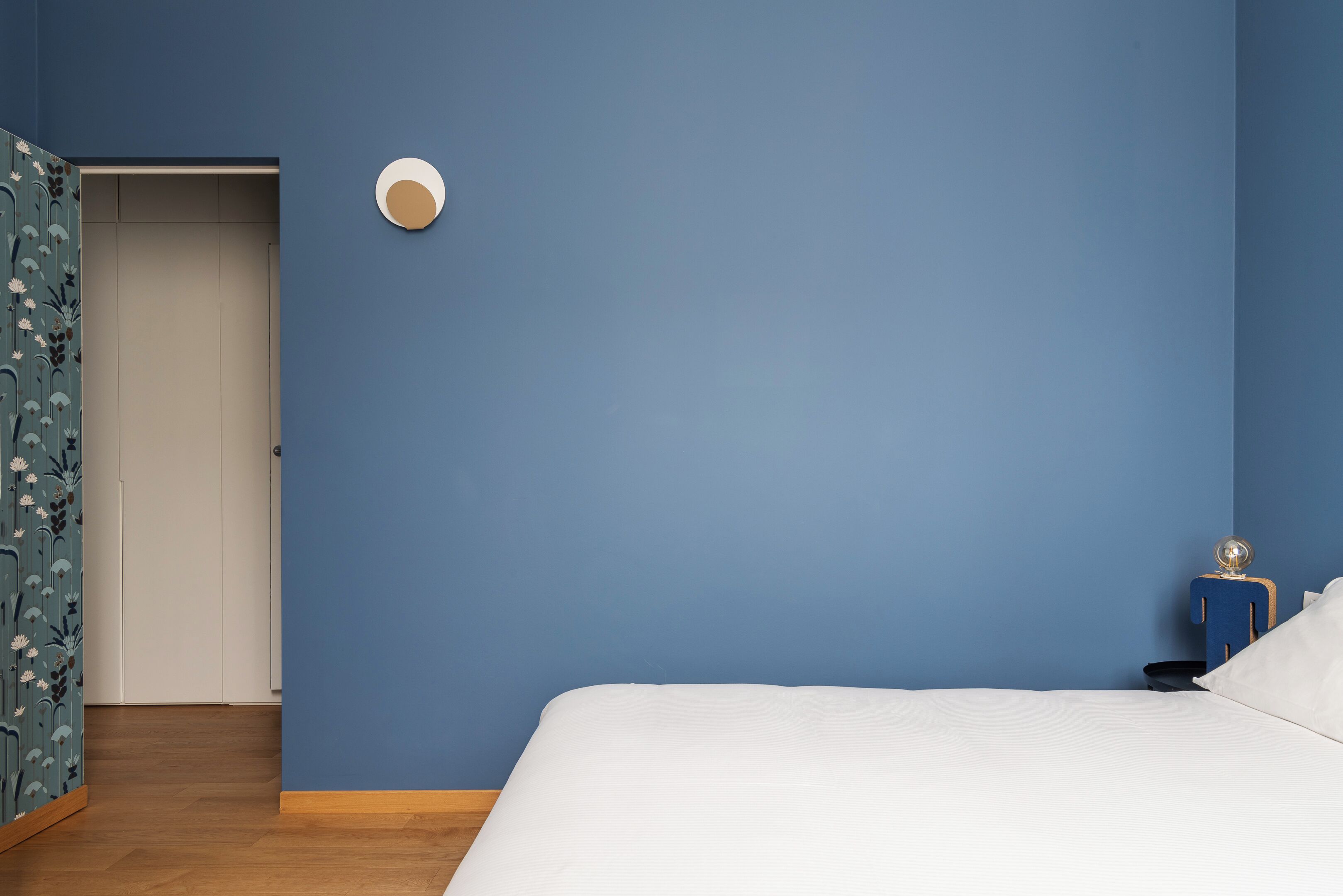 A bedroom with a blue wall, a white bed, a wooden nightstand with a lamp, and a door with floral wallpaper.