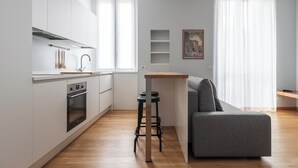 Una cocina moderna con armarios blancos, horno integrado y encimera de madera. Hay un sofá gris y una pequeña mesa de madera con taburetes.