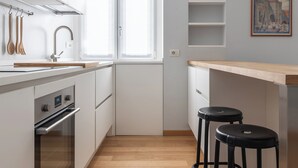 Una cocina moderna con armarios blancos, encimera de madera y horno empotrado. Hay dos taburetes negros y una ventana con cortinas blancas.
