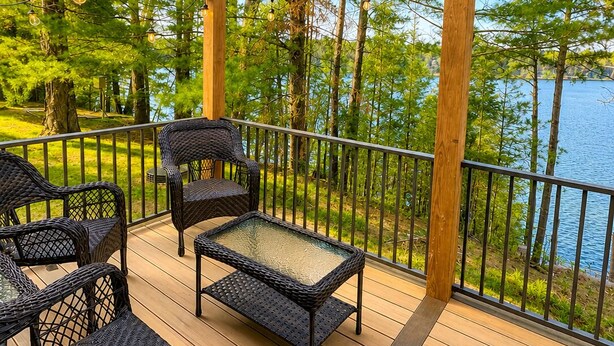 Una terraza de madera con mobiliario de mimbre y luces colgantes, con vistas a un cuerpo de agua y árboles.