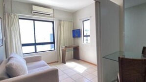 A modern living room with a sofa, a glass table, and a chair. There is a TV mounted on the wall, and a window with curtains.