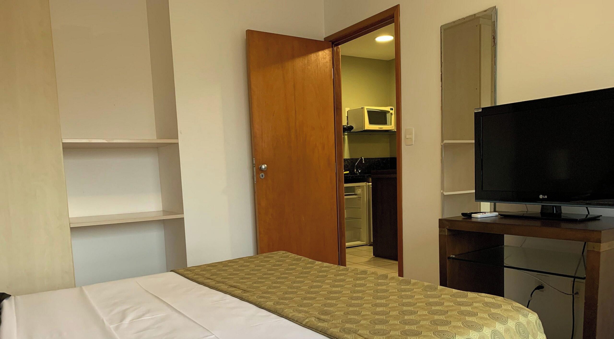Un dormitorio con una cama, televisión, armario y una puerta que da a la cocina.