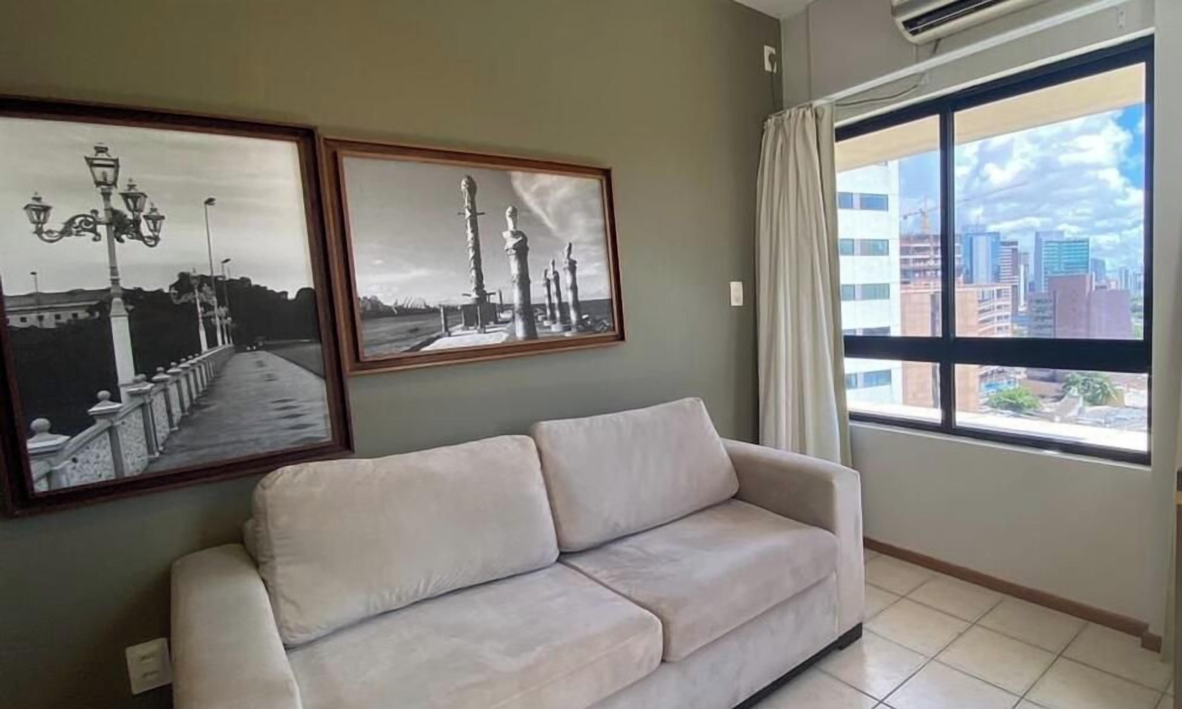 A room with a sofa, two framed pictures on the wall, a large window with a view of buildings, and an air conditioning unit.