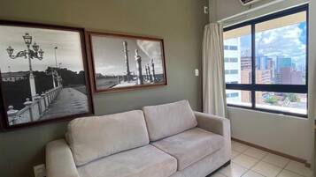 A room with a sofa, two framed pictures on the wall, a large window with a view of buildings, and an air conditioning unit.